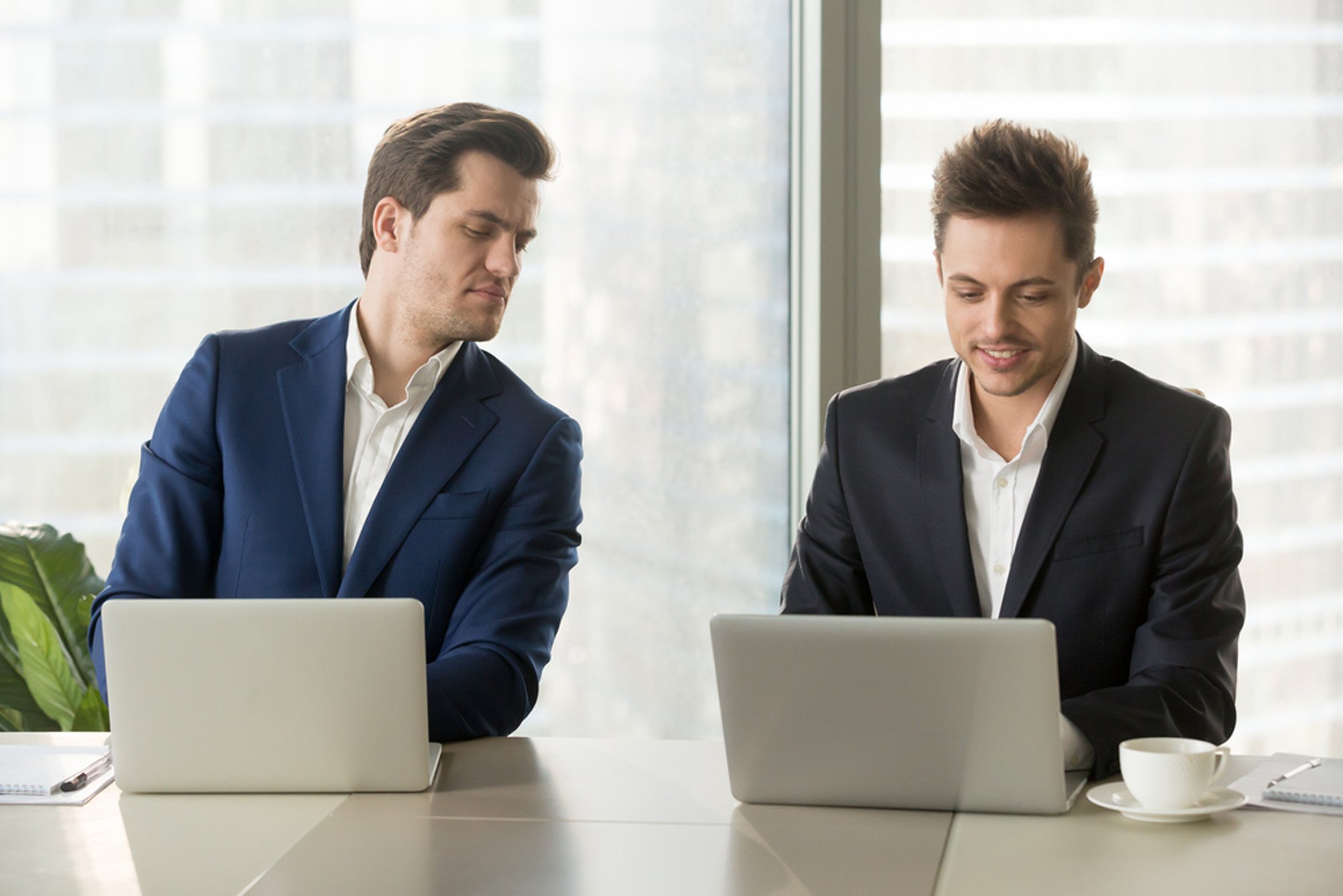 Two men in business suits are looking at their laptops, one is looking over at his friend's laptop