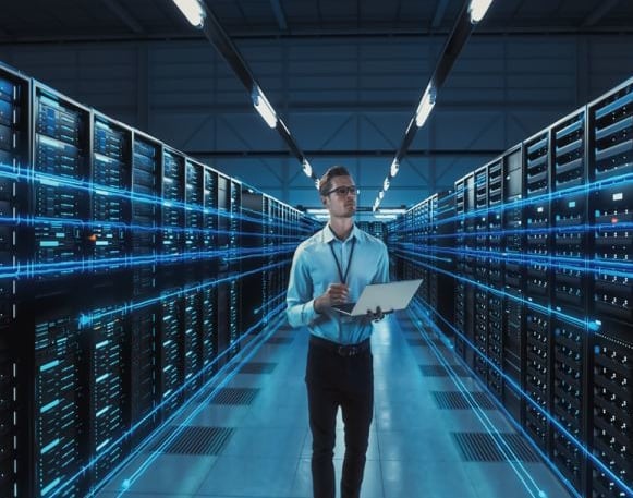 Man in a blue button-up shirt stands in a server room holding an open laptop