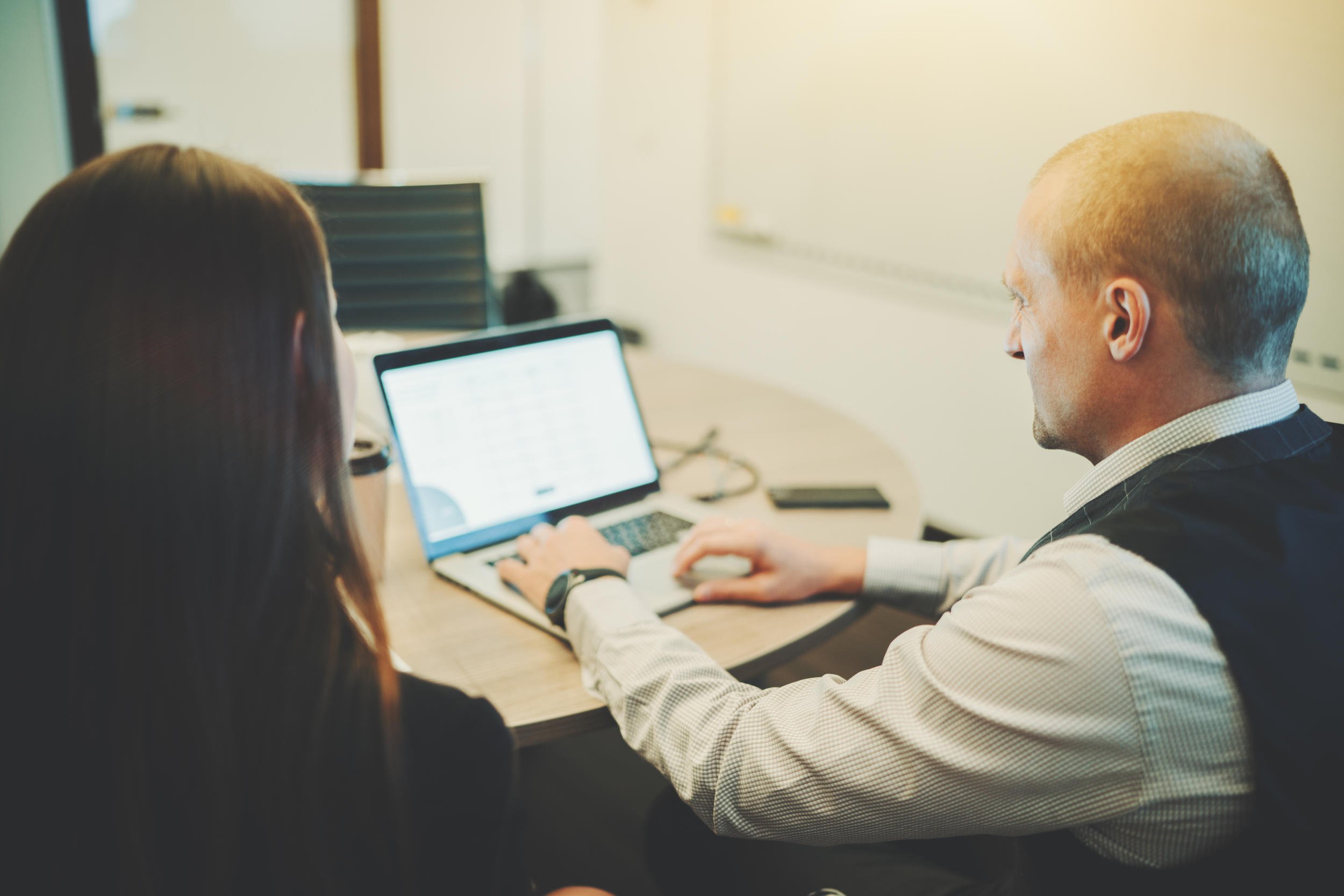 Older man shows something to a woman on a laptop, both are facing away from the camera