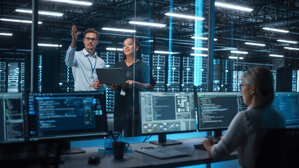 Two IT specialists with a laptop discuss server racks in a data center while a colleague monitors dashboards