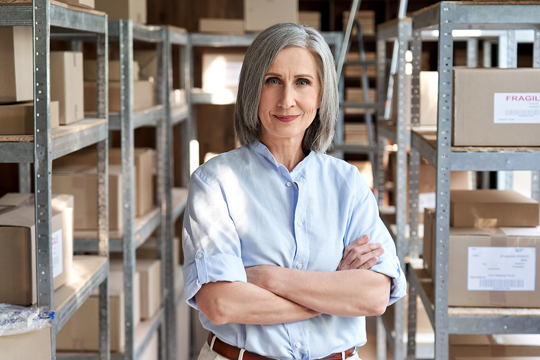 Confident older woman with grey hair stands in a storage room with her arms crossed, half smiling at the camera