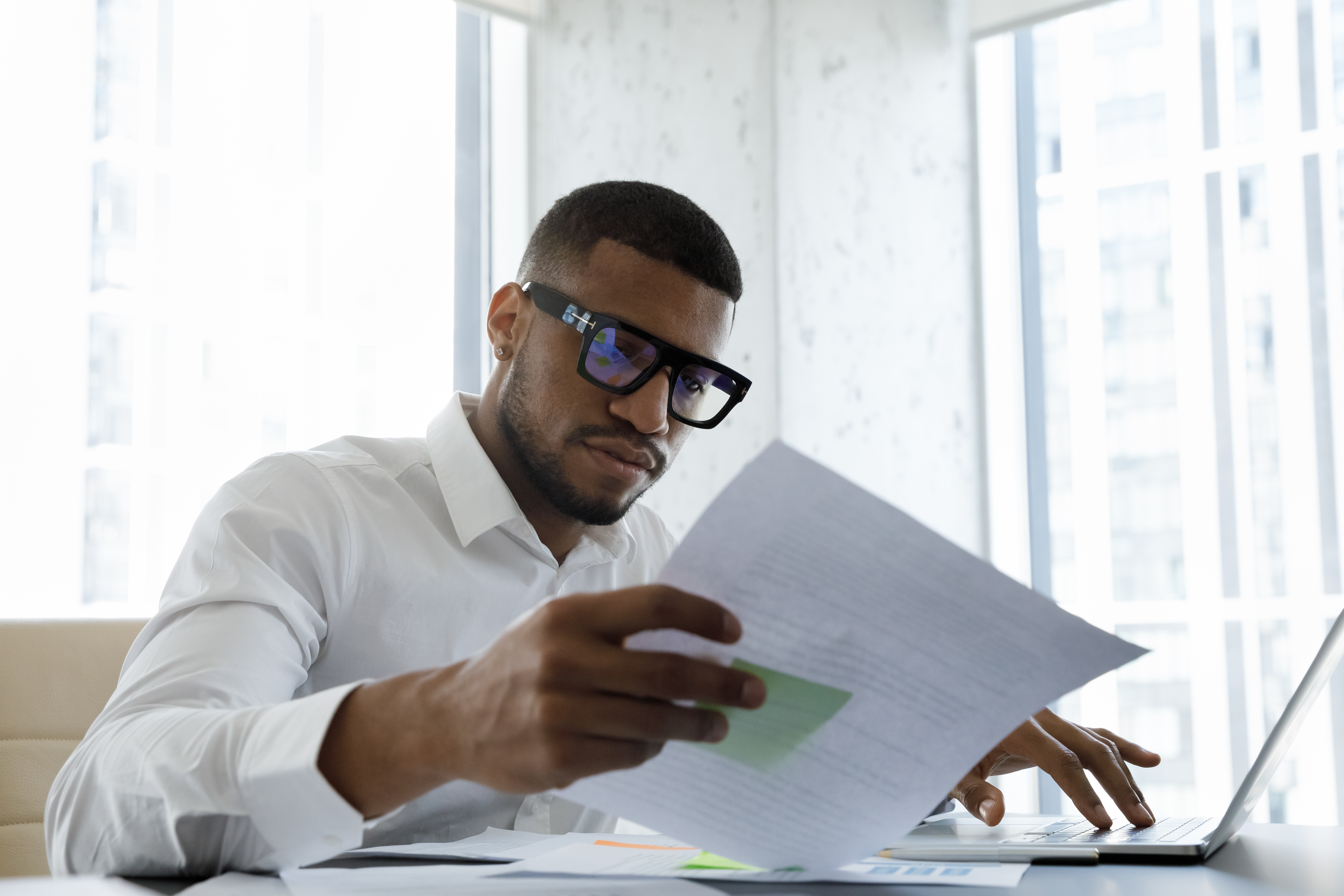 Man with dark-frame glasses looking at some papers in his hand while he does something on a laptop with the other