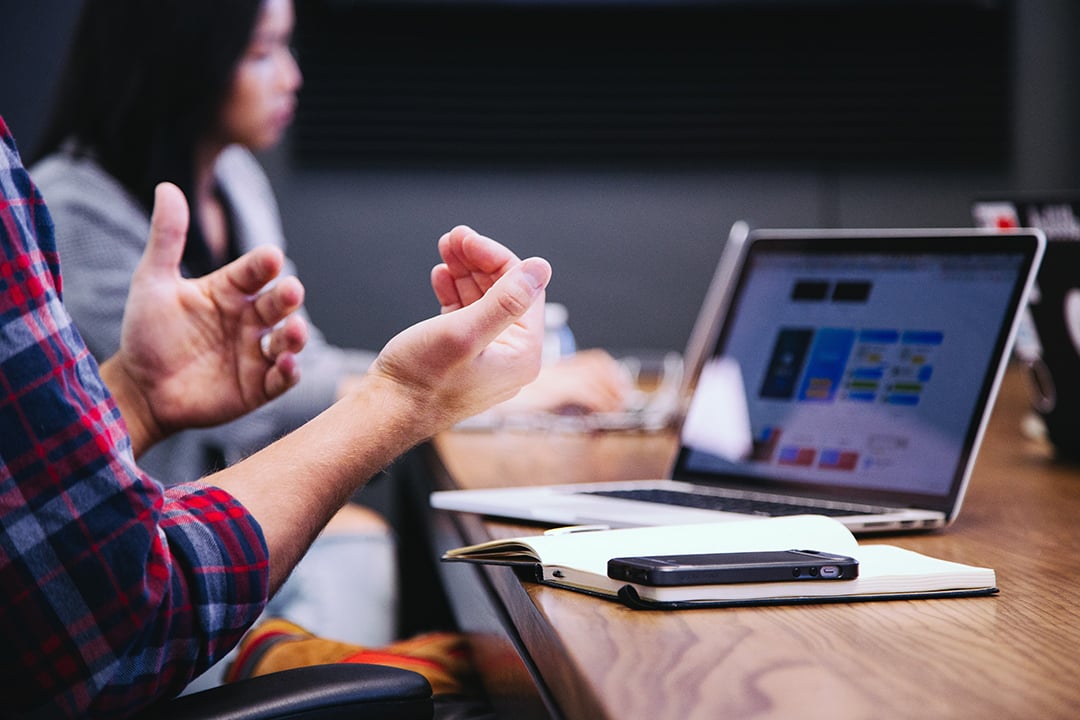 A man's hands are expressive while he speaks, his laptop is visible in front of him.