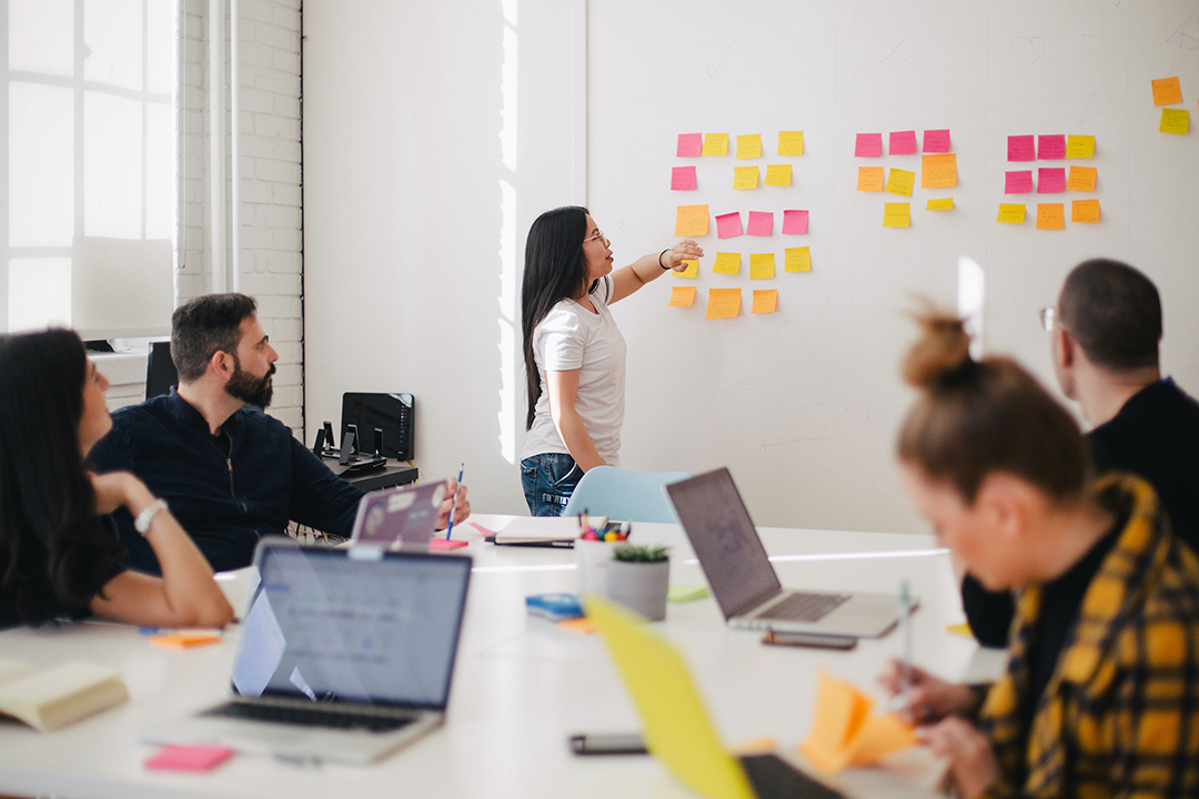 Several people sit around a table in an office setting, one woman is putting post-its on a whiteboard